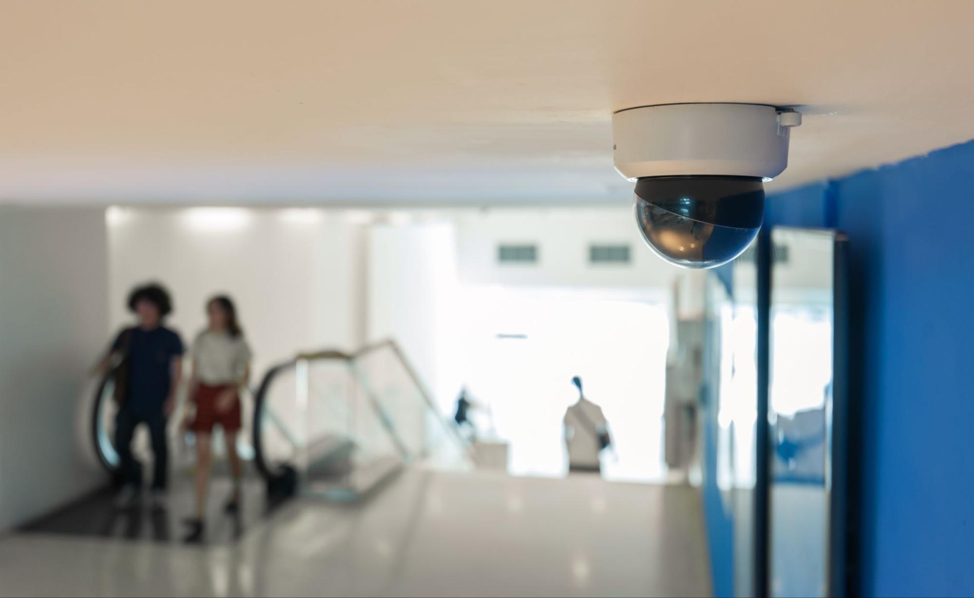 A dome-style Smart CCTV Camera mounted on a ceiling overlooks a modern shopping mall or office lobby with ran escalato