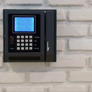 A wall-mounted black key card reader and keypad, characteristic of Modern Intercom Systems or time and attendance devices, set against a rough white brick wall
