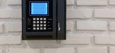 A wall-mounted black key card reader and keypad, characteristic of Modern Intercom Systems or time and attendance devices, set against a rough white brick wall